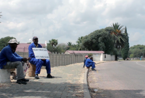Black workers in blue overalls waiting in a road side with a sign reading electrician and a phone number