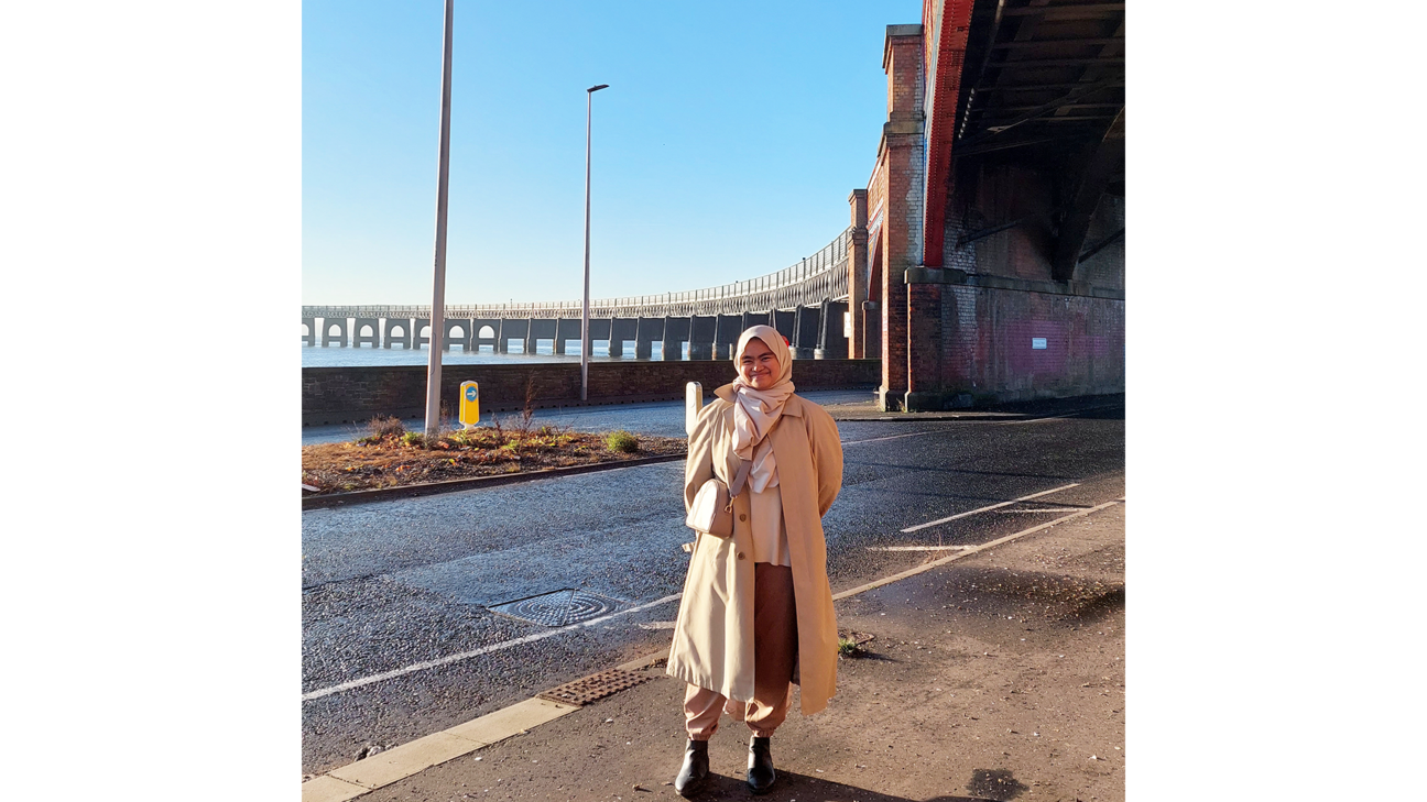 Azizah Azis standing under the Tay Rail Bridge with the River Tay behind her