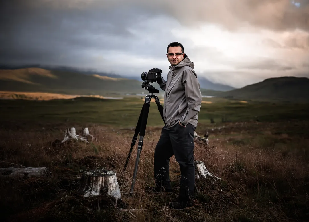 Shahbaz standing with his camera on a tripod with hills behind him