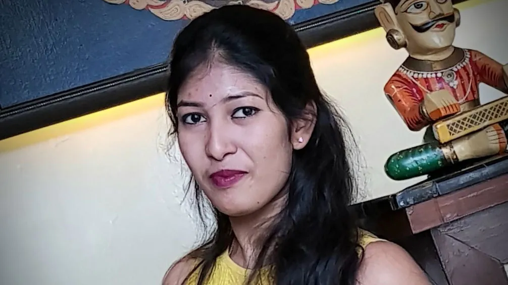 Photo of a graduate, Nandhni Ghandi standing in front of a cabinet with a stature of a man sitting cross legged