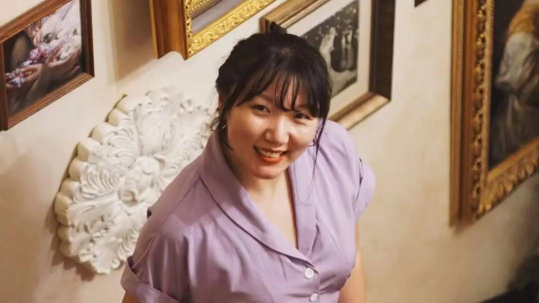 Jiang Hong stands on the stairs with ornate paintings on the wall behind her.