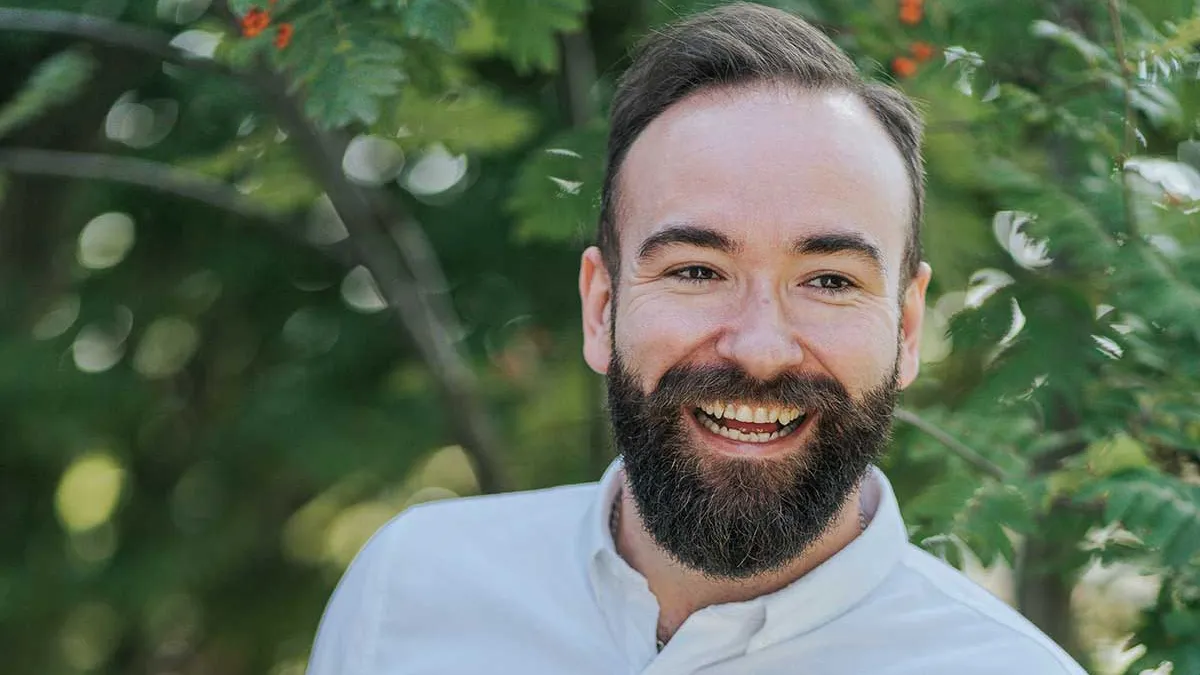 A man with dark brown hair and a beard smiling in front of a tree with berries