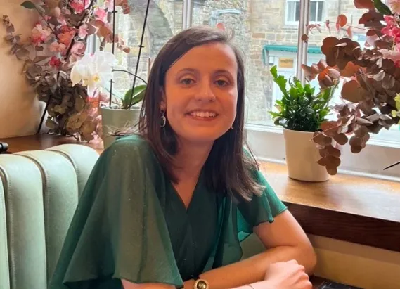 A woman sitting on an upholstered bench leaning her arms on a table in front. There are plants in pots in a window behind her.