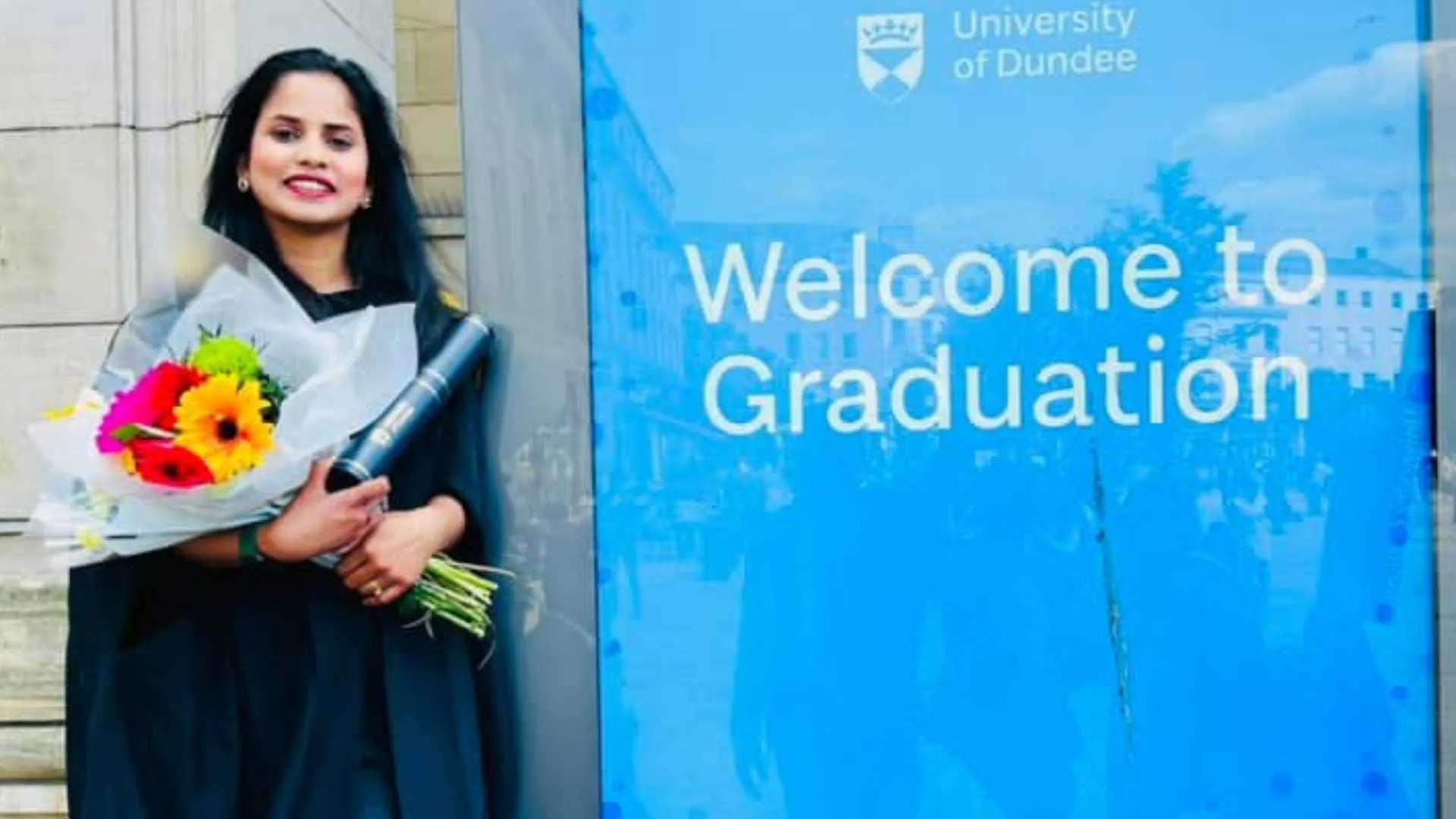 Thusharika holds a bouquet of flowers, leaning against a building and wearing graduation robes. There is a large blue sign saying "Welcome to graduation"