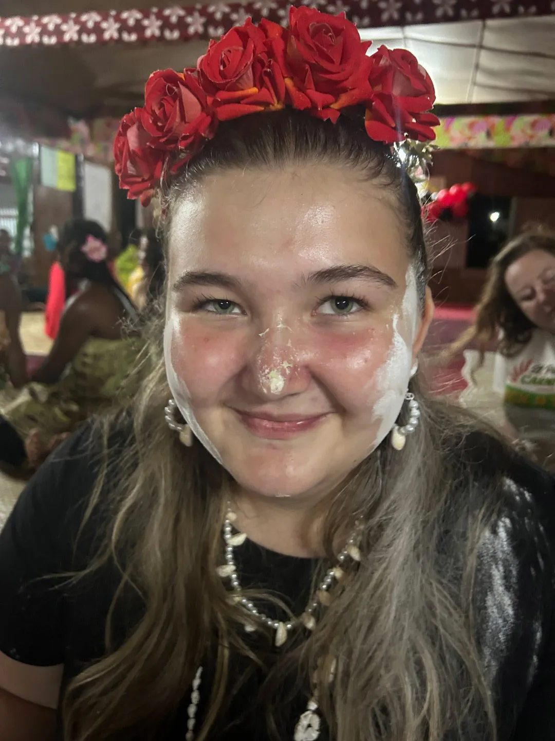 A close-up of a young woman wearing a red rose crown, shell jewellery, and white ceremonial powder on her face. Other people sit on the woven mat behind her.