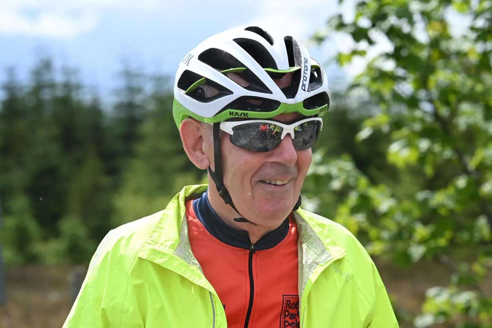 A smiling cyclist wearing a bright yellow jacket, white helmet, and sunglasses, standing outdoors.