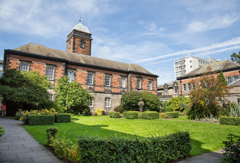 a red stone building with grass, seating and hedges out the front
