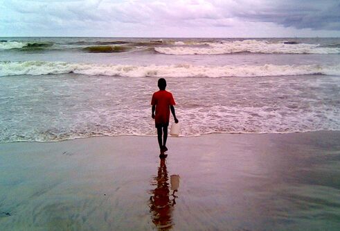 A boy is walking toward the shoreline on a beach, with a white bucket in his hand