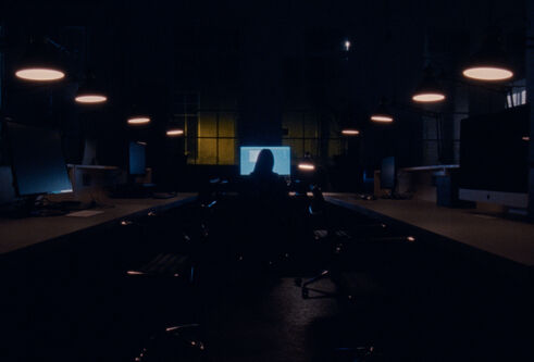 Film still from ‘The Dead Are Jewels To Me’ – image of Dr Maria Maclennan taken from behind sitting in the distance watching a screen in a darkened room.