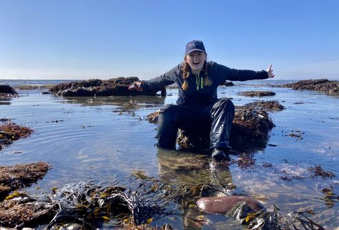 a young woman in her 20's surrounded by rockpools of shimmering silver and blue sea water on a clear, blue skied day