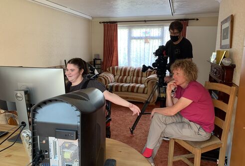 a middle aged man sat on wooden chair, looking with interest at a young, late-teened girl sat in a wheelchair as she works on a computer