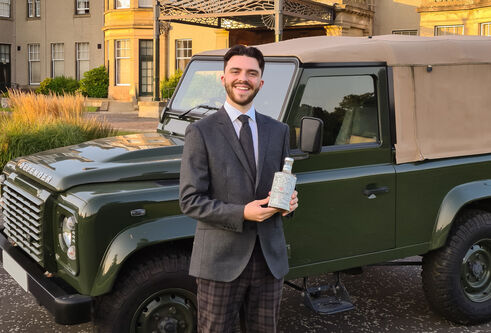 Lewis Kenned holds a bottle of his non-alcoholic gin with a Land Rover and traditional Scottish country hotel behind him