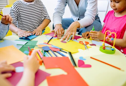 An adult and group of children around a desk that is covered in brightly coloured paper and crayons