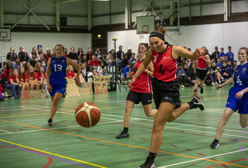 a group of young women in their 20's, some wearing red sporting gear and others blue, running on a basketball pitch chasing a ball