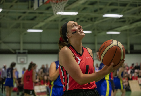 young woman in her 20's wearing red sporting tank top, holding orange basketball with one hand 