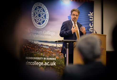 middle aged man standing on stage behind podium, wearing dark navy suit and speaking to a crowd of people who can be seen in foreground