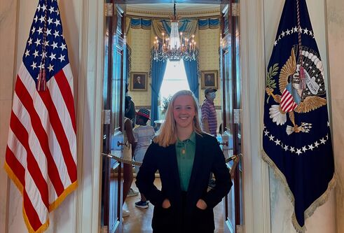 Young girl in her early 20's wearing a black suit jacket stands posing in front of an open door to the Oval Office, which is the office of the President of the USA. On her left a large American flag hangs down and on her right is the President Seal flag