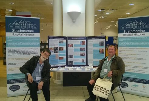 Two community members sit in front of a stall advertising the work of the Strathmartine Hospital Histories project.