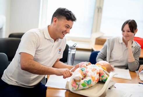 A male and female, both in Dundee University branded shirts are sitting at a table with a baby doll between them.