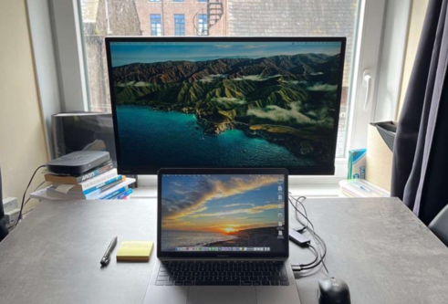 A laptop and computer monitor sit on a desk in front of a window. There is a pen and sticky notes sitting to the left on the desk.