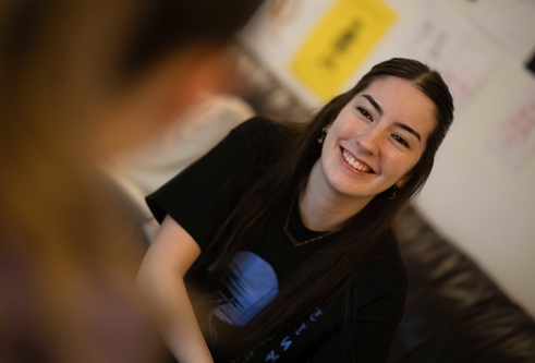 A girl with long dark hair smiles towards a person who is blurred in the foreground.