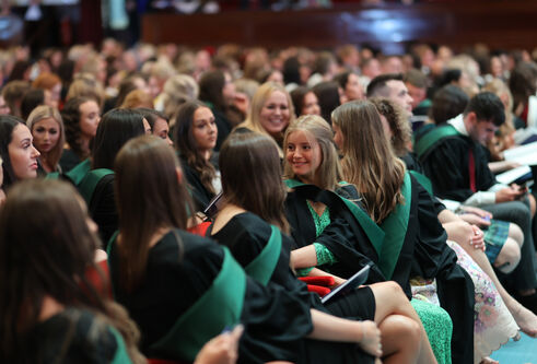 A group of students in black graduation robes with green hoods sitting in a theatre smiling at eachother