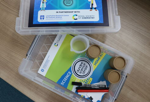 Storage box and lid on a wooden table with science equipment and a green and blue leaflet inside.