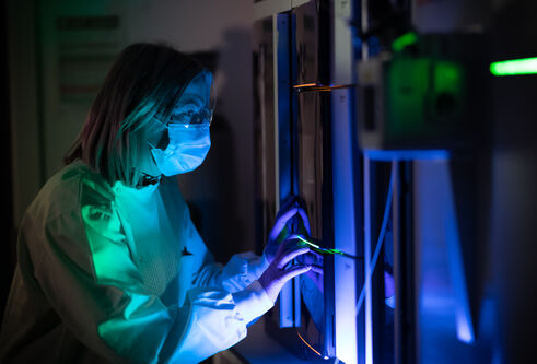 a women in a lab coat and mask is lit with blue and green lab as she does work in the lab