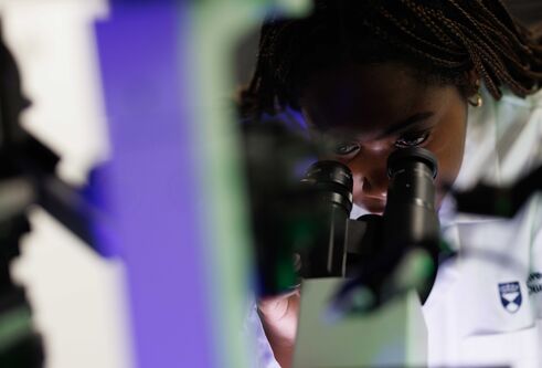 A woman in a lab coat looks through a microscope. A blue-green line cuts across the image.