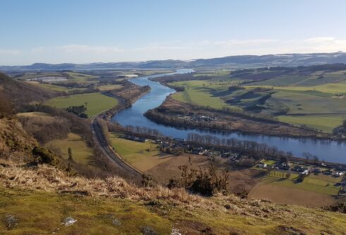 Landcape shot taken high up a hill, showing a river streaming though hills and fields and into the far distance