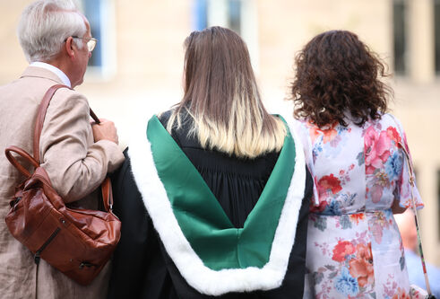 Three people standing with their back to the camera. Young female in centre wearing a graduation gown and green hood lined with white fur. Older male on left holding brown bag over his shoulder and older woman on first hand side wearing floral dress.