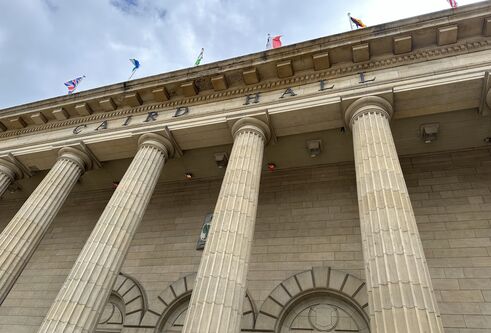 Roof area of old building with tall pillars and flags out of the top, and a cloudy blue sky above