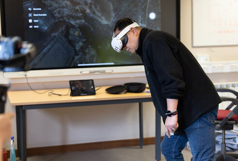 Man wearing virtual reality headset bending to side with screen behind him in classroom setting
