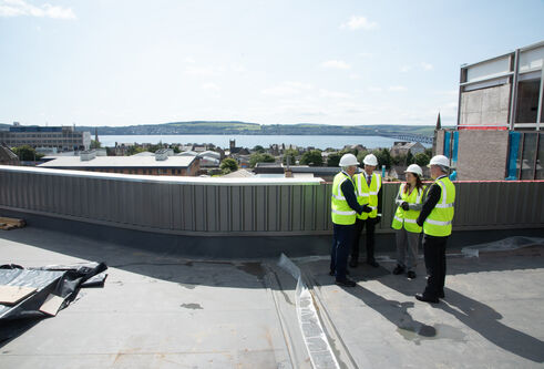 A group of people in high vis vests are stood on a roof in a group speaking to eachother. There is a beautiful backdrop showing the river tay behind them