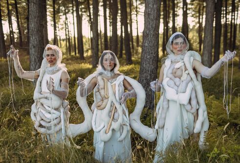Three women wearing 3D fabric body parts representing the human gut on their outfits with their arms up dancing in a forest 