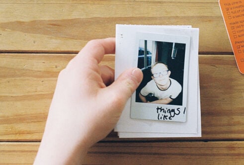 A hand holds a stack of cards with a polaroid photo on top against a wooden background. In the upper right hand corner is an orange card with a number of short lists typed on it. There are tickboxes against each item on the lists