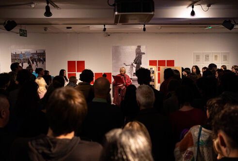 A group of people watching a woman speak in front of an art gallery exhibition