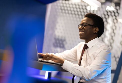 A man in a white shirt and black tie sits at a laptop, smiling.