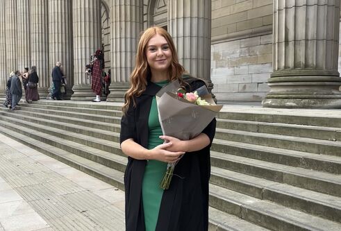 A person stands on the steps outside of a building with large pillars, they are wearing graduation robes and a green dress, holding a bunch of flowers.