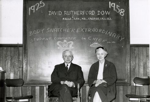 A black and white photo of Professor David Dow and Dr Agnes Dow sitting in front of a blackboard. The board reads 1925 1958 David Rutherford Dow Body Snatcher Extraordinary Student Comforter in Chief etc., etc. Above David's head is a crown drawn on the blackboard, above Agnes's, a tiara.