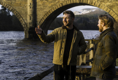 Man standing next to river, gesturing, with bridge in the background