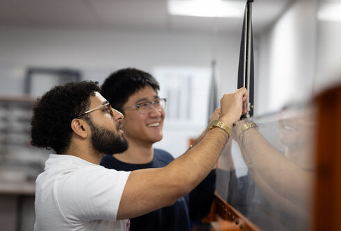 Two people stand together in front of a large piece of glass, one of them holds a device against it.