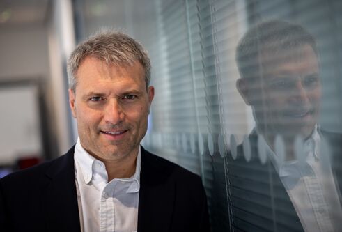 Alessio Ciulli in black jacket, leaning against a glass window, with blinds closed behind it.