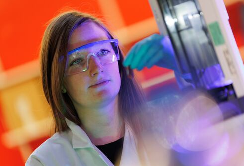 Head and shoulders shot of woman with medium length dark hair, wearing safety glasses and a lab coat, looking at a computer screen