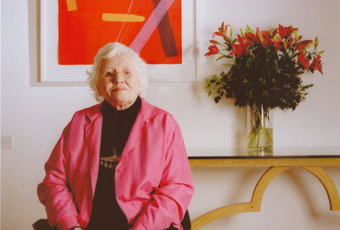 An older woman with white hair sits indoors wearing a bright pink jacket, with an abstract red and orange artwork framed on the wall behind her and a vase of red lilies on a table to her side.