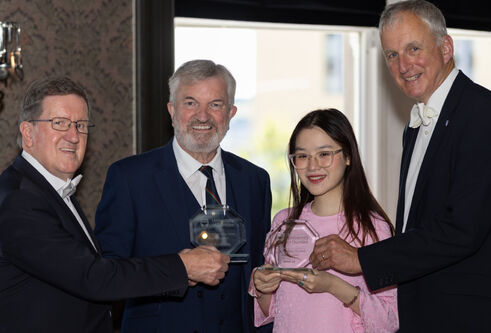 Four people smiling and holding clear glass Bridge Alumni Awards. From left to right: George Robertson, John Fyffe MBE, Heidi Lee, and Nigel Seaton. The group stands indoors with soft lighting and patterned wallpaper in the background.