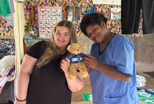 Two people standing indoors on a woven mat, smiling and holding a University of Dundee teddy bear together. Colourful fabric and hanging clothes are visible in the background.