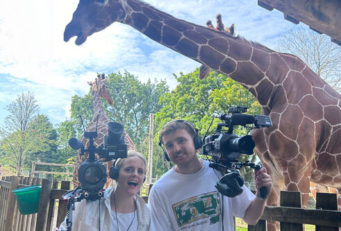Graduate Euan McClenaghan poses with a colleague and their cameras in front of a giraffe