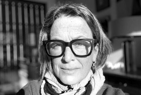 A black-and-white close-up portrait of a person wearing large square glasses, looking toward the camera with bright sunlight casting strong shadows across their face.