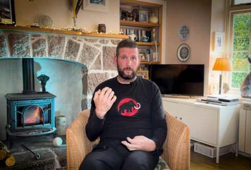 man sitting on sofa, holding hands up, in living room with wood burning fire behind